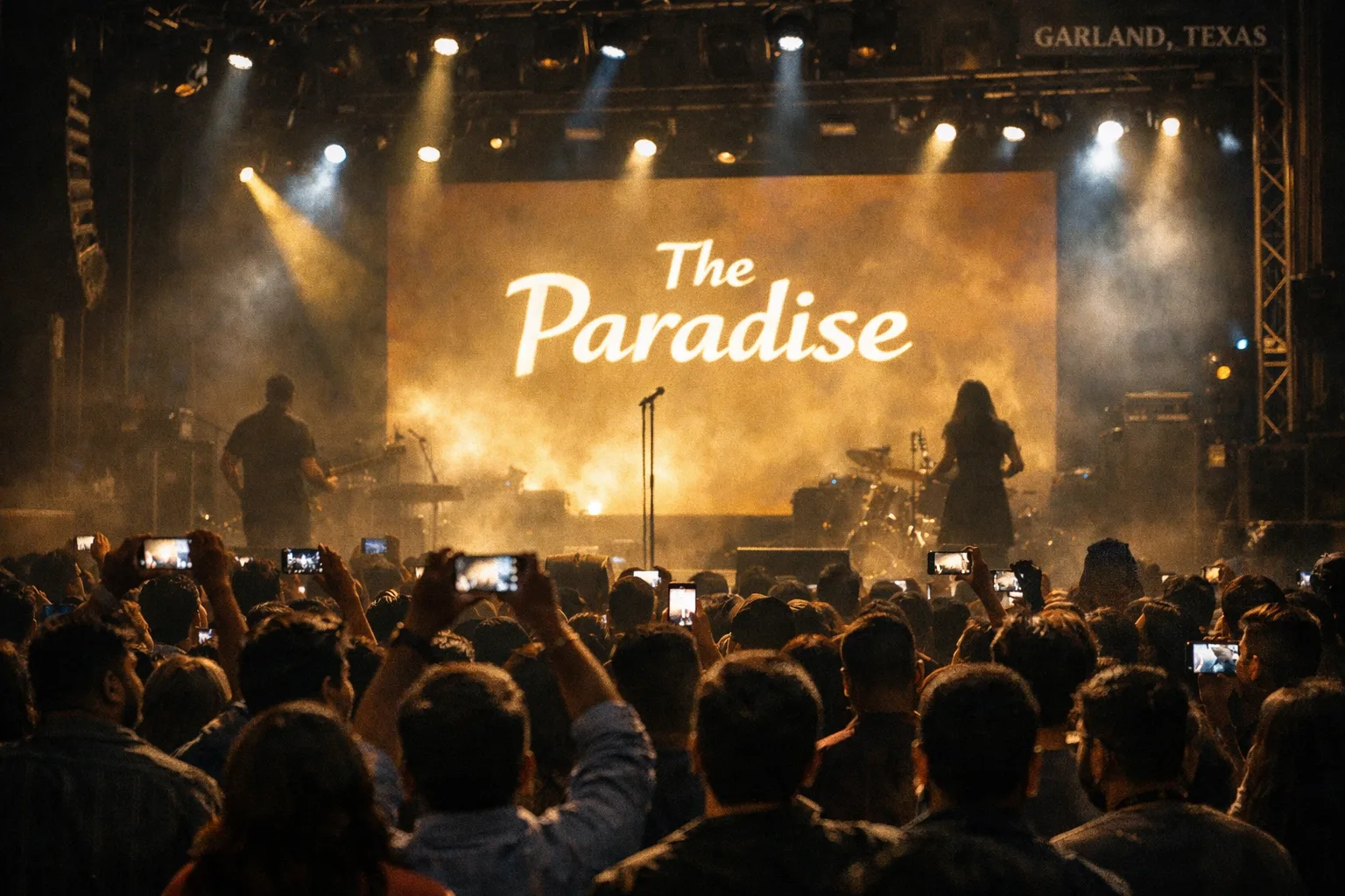 A packed indoor concert venue in Garland with stage lights, a film-promotion screen and indistinct audience members holding phones.